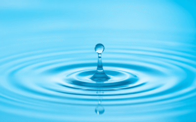 Közeli kép egy tiszta vízcseppről, amely hullámokat kelt a nyugodt vízen, lágyan fénylő visszatükröződésekkel egy békés kék háttér előtt.